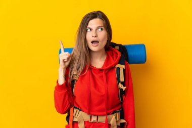 Sarı arka planda izole edilmiş büyük bir sırt çantası olan Slovak dağcı kadın parmağını yukarı kaldıran bir fikir düşünüyor.