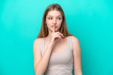 Genç Rus kız mavi arka planda izole edilmiş. Sessiz bir jest işareti var. Parmağını ağzına sokuyor.