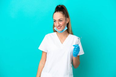 Beyaz dişçi kadın, elinde mavi arka planda izole edilmiş aletlerle yan tarafa bakıyor ve gülümsüyor.