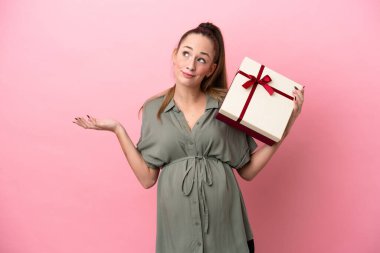 Pembe arka planda izole edilmiş genç bir kadın hamile ve elinde şüpheleri varken bir hediye tutuyor.