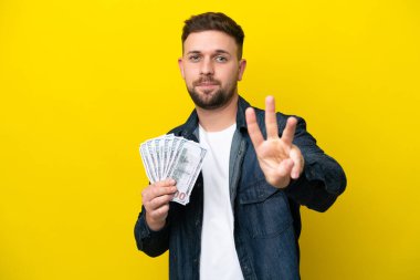 Sarı arka planda izole edilmiş bir sürü para alan genç beyaz adam mutlu ve parmaklarıyla üçe kadar sayıyor.