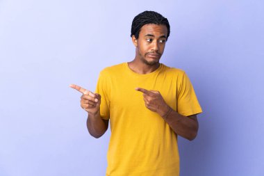 Örgülü genç Afrikalı Amerikalı adam mor arka planda izole edilmiş korkmuş ve bir tarafı işaret ediyor.