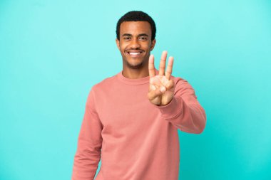 Mavi arka planda yaşayan Afro-Amerikalı yakışıklı adam mutlu ve parmaklarıyla üçe kadar sayıyor.