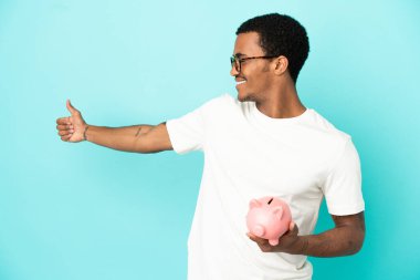 Afro-Amerikalı yakışıklı bir adam kumbarasını izole edilmiş mavi arka planda tutuyor ve baş parmak işareti yapıyor.