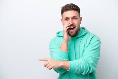 Young caucasian man isolated on white background pointing to the side to present a product and whispering something