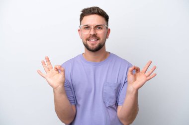 Beyaz arka planda izole edilmiş genç beyaz bir adam iki eliyle tamam işareti gösteriyor.