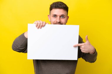 Young caucasian man isolated on yellow background holding an empty placard and hiding behind it
