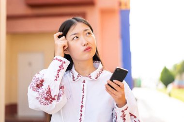 Genç Çinli bir kadın açık havada cep telefonu kullanıyor. Şüpheleri var ve kafası karışık.