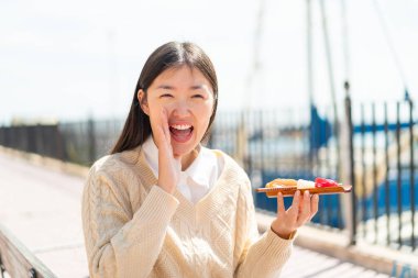 Genç Çinli bir kadın açık havada sashimi tutuyor ve ağzı açık bağırıyor.