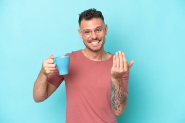 Kafkasyalı genç bir adam mavi arka planda izole edilmiş bir fincan kahve tutuyor ve elini uzatmaya davet ediyor. Gelmene sevindim.