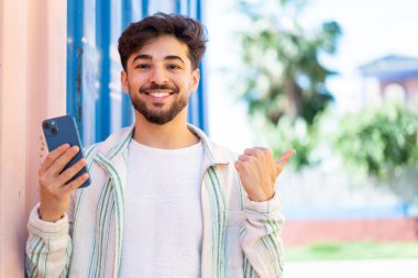 Yakışıklı bir Arap, açık havada cep telefonu kullanıyor. Bir ürünü sunmak için yan tarafı işaret ediyor.