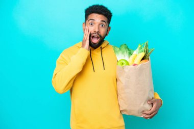 Genç Brezilyalı bir adam mavi arka planda izole edilmiş bir paket yiyecek alıyor. Şaşırmış ve şok olmuş bir yüz ifadesi var.