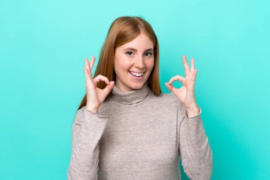 Mavi arka planda izole edilmiş genç kızıl saçlı kadın iki eliyle onay işareti gösteriyor.