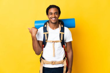 Sarı arka planda izole edilmiş büyük bir sırt çantalı genç dağcı bir ürünü sunmak için kenarı işaret ediyor.