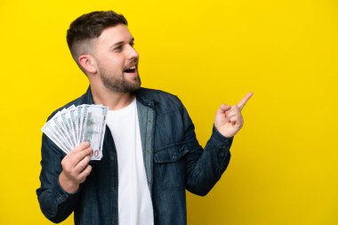 Genç beyaz adam sarı arka planda izole edilmiş bir sürü para alıyor. Çözümü bulmak için parmağını kaldırıyor.