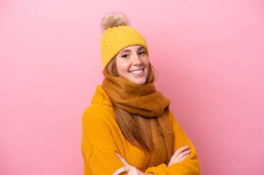 Kışlık ceket giyen genç kızıl saçlı kadın pembe arka planda izole edilmiş, kolları bağlı ve ileriye bakıyor.