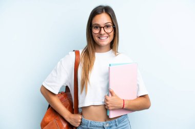 Mavi arka planda izole edilmiş genç bir bayan kolları kalçasında ve gülümsüyor.