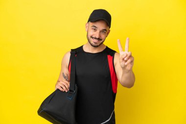Sarı arka planda izole edilmiş spor çantalı genç beyaz adam gülümsüyor ve zafer işareti gösteriyor.