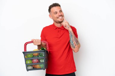 Beyaz arka planda izole edilmiş yiyecek dolu bir sepet taşıyan genç beyaz bir adam yukarı bakarken bir fikir düşünüyor.