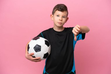 Kafkas futbolcu çocuk pembe arka planda izole edilmiş, olumsuz bir ifadeyle baş parmağını aşağı gösteriyor.