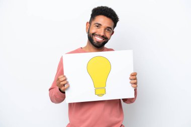 Genç Brezilyalı bir adam beyaz arka planda izole edilmiş elinde mutlu ifadeli bir ampul simgesi olan bir levha tutuyor.
