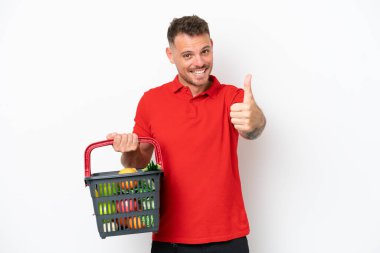 Beyaz arka planda izole edilmiş yiyecek dolu bir sepet taşıyan beyaz bir adam. Başparmakları havada, çünkü iyi bir şey oldu.