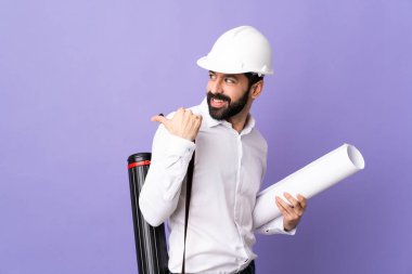 Miğferli genç bir mimar, elinde bir ürün sunmak için kenarı işaret eden izole edilmiş mor arka plan planları var.