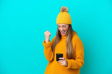 Kışlık ceket giyen genç beyaz kadın mavi arka planda izole edilmiş ve telefon zafer pozisyonunda.