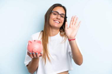 Elinde kumbara tutan beyaz bir kadın mavi arka planda izole edilmiş mutlu bir ifadeyle selam veriyor.