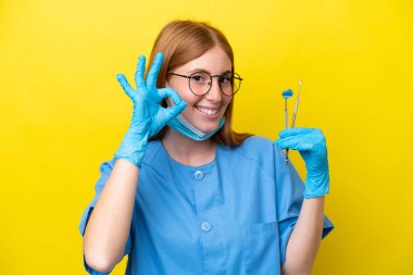 Genç kızıl saçlı dişçi kadın sarı arka planda izole edilmiş. Parmaklarıyla tamam işareti gösteriyor.