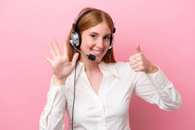 Tele pazarlamacı kızıl saçlı kadın pembe arka planda izole edilmiş kulaklıkla çalışıyor. Parmaklarıyla altı kişi sayıyor.