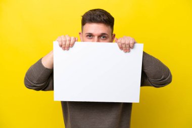 Young caucasian man isolated on yellow background holding an empty placard and hiding behind it