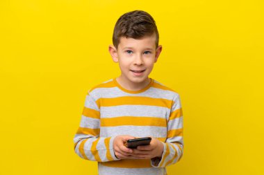 Sarı arka planda izole edilmiş küçük beyaz çocuk cep telefonuyla bir mesaj gönderiyor.