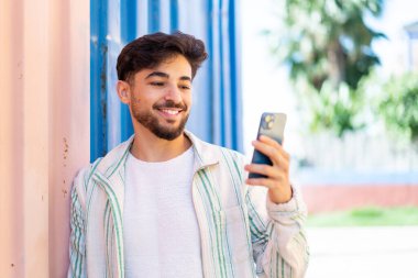 Yakışıklı bir Arap açık havada mutlu bir ifadeyle cep telefonu kullanıyor.