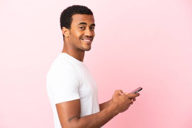 İzole edilmiş pembe arka plandaki Afrikalı yakışıklı adam cep telefonuyla mesaj ya da e-posta yolluyor.
