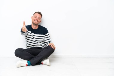 Genç adam beyaz arka planda izole bir şekilde yerde oturuyor ve iyi bir anlaşma için el sıkışıyor.