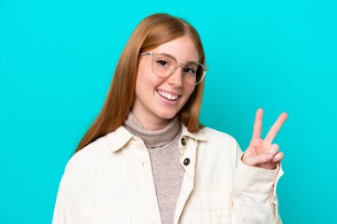 Genç kızıl saçlı kadın mavi arka planda izole edilmiş gözlüklü ve iyi işaretiyle