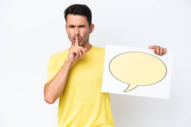 İzole edilmiş beyaz arka plan üzerinde genç yakışıklı bir adam elinde konuşma baloncuğu ikonlu bir levha ile sessizlik hareketi yapıyor.