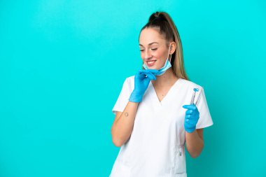 Beyaz dişçi kadın, elinde mavi arka planda izole edilmiş aletlerle yan tarafa bakıyor ve gülümsüyor.