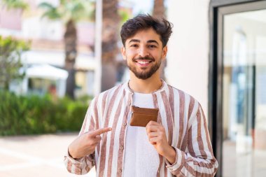 Yakışıklı bir Arap, dışarıda bir cüzdan tutuyor ve ona doğrultuyor.