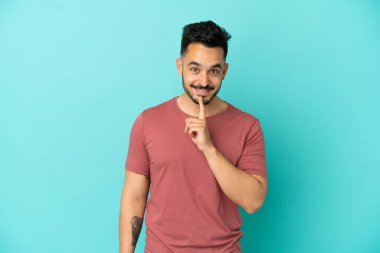 Mavi arka planda izole edilmiş genç beyaz adam sessiz bir jest işareti gösteriyor. Parmağını ağzına sokuyor.