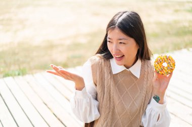 Genç Çinli bir kadın açık havada yüzünde sürpriz bir ifadeyle donut tutuyor.