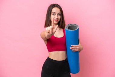 Genç sporcu Ukraynalı kadın yoga derslerine gidiyor. Elinde pembe arka planda izole edilmiş bir minder var. Parmağını kaldırıp kaldırıyor.