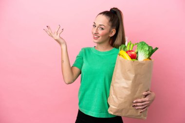Alışveriş torbası tutan genç bir kadın. Davet etmek için ellerini yana uzatıyor.