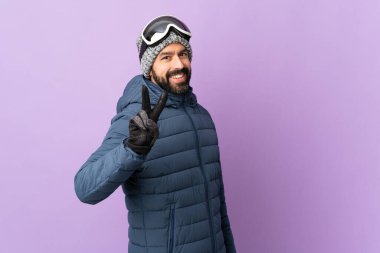 İzole edilmiş mor arka planda snowboard gözlüklü kayakçı gülümseyip zafer işareti gösteriyor.