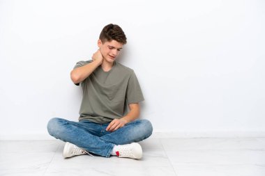 Genç Rus bir adam beyaz arka planda boynu ağrıyan bir şekilde yerde oturuyor.
