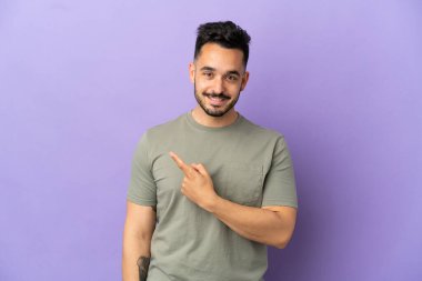 Mor arka planda izole edilmiş genç beyaz adam bir ürünü sunmak için kenarı işaret ediyor.