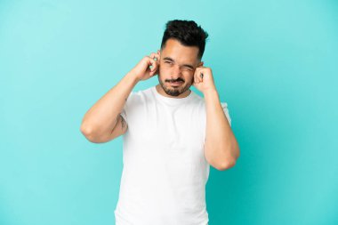 Mavi arka planda izole edilmiş genç beyaz adam hayal kırıklığına uğramış ve kulaklarını kapatmış.