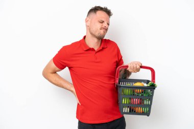 Beyaz arka planda bir sepet dolusu yiyecek taşıyan beyaz tenli genç bir adam gayret gösterdiği için sırt ağrısından muzdarip.