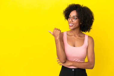 Sarı arka planda izole edilmiş genç Afrikalı Amerikalı kadın bir ürünü sunmak için kenarı işaret ediyor.